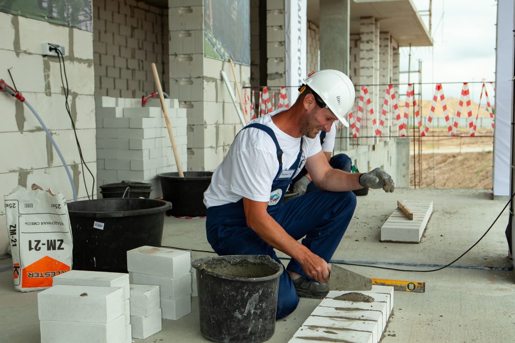 A man in white hard hat and blue overalls working on a building.